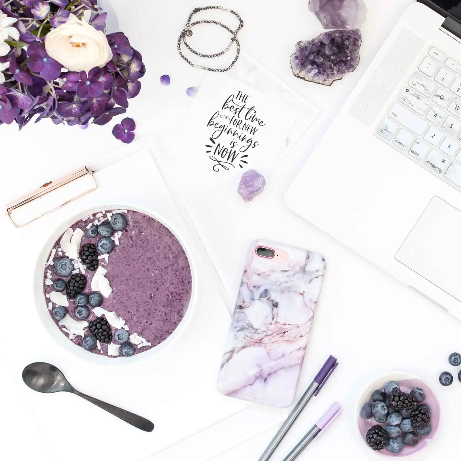 Flat lay photo of a white surface with white and purple objects, such as an amethyst rock, purple flowers, a purple smoothie bowl, a purple marble print phone case, a silver laptop, and purple pens, artfully arranged.