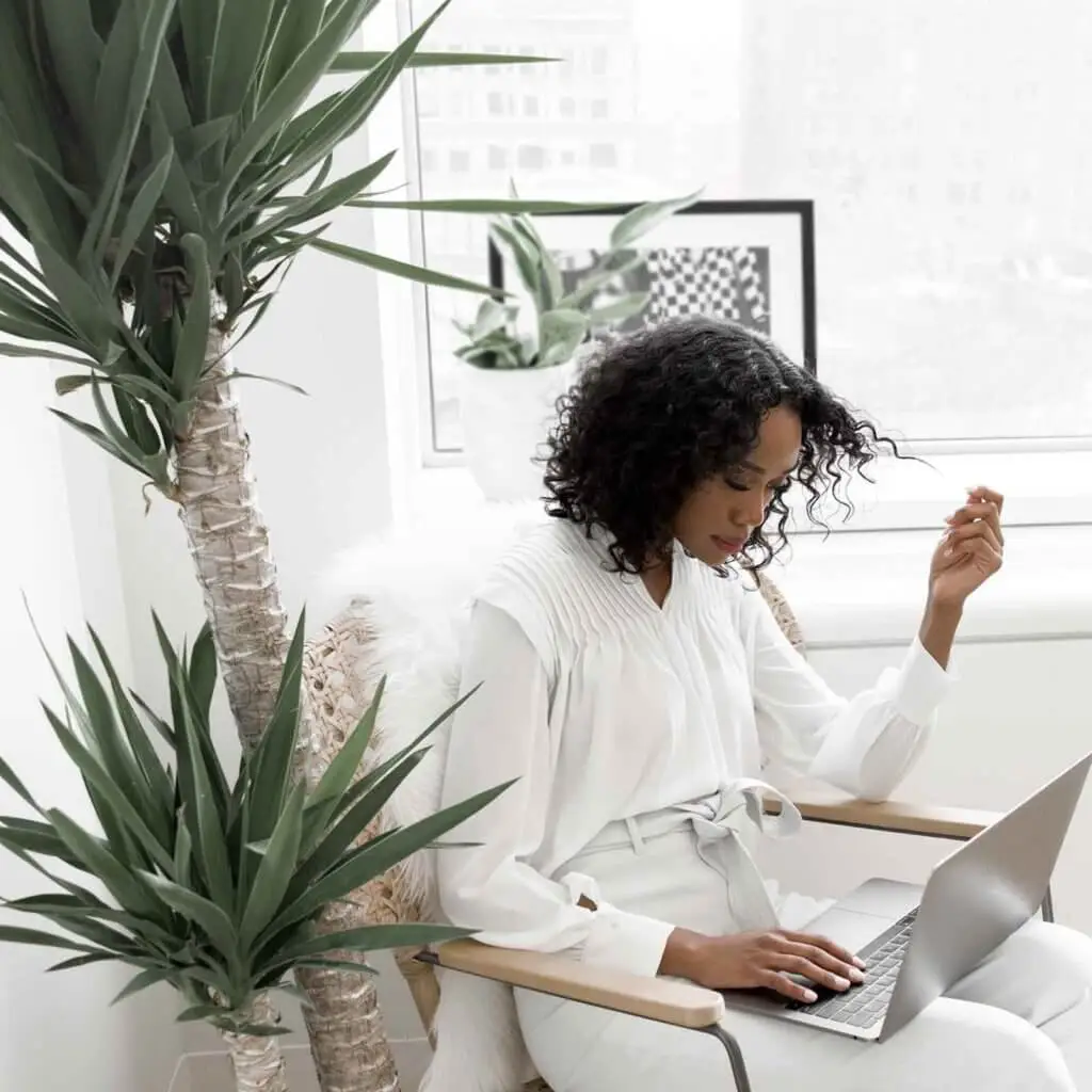 Photo of a woman wearing a white blouse and light grey pants reading something on her laptop while twirling her curly hair with one hand.