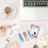 Flat lay photo of a light pink surface with artfully arranged items such as pastel printed photos, a silver laptop, a white rose, and a cup of espresso with a macaron on the side.