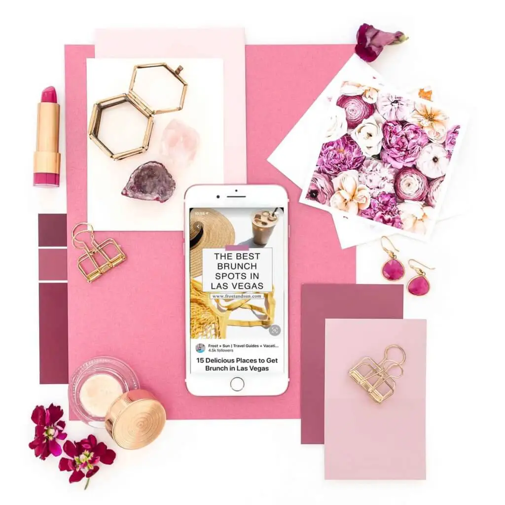 Flat lay of pink and gold objects, such as lipstick, papers, flowers, and makeup, artfully arranged. In the middle is a white iPhone open to a pin on Pinterest with extra data like profile photo, profile name, and blog post title.