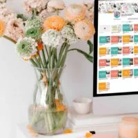 Closeup photo of a white desk with pink and peach office accessories, a clear vase full of pink, peach, and white flowers, and a Mac computer open to a Pinterest profile page.