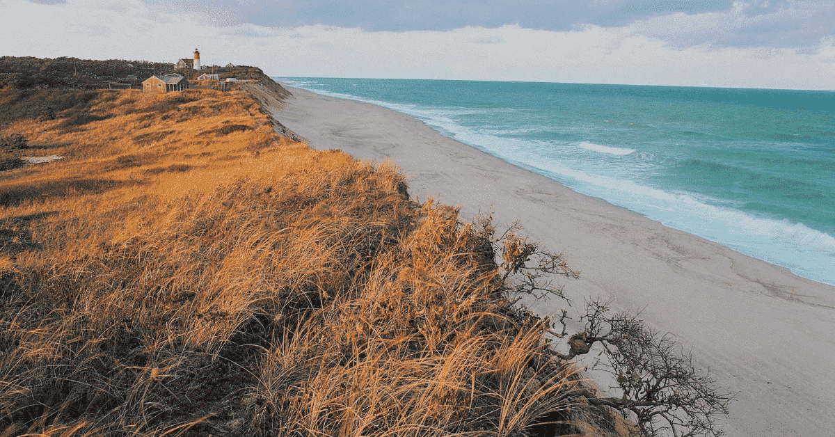 Landscape photo of a beach and sand dunes with a lighthouse in the far distance.