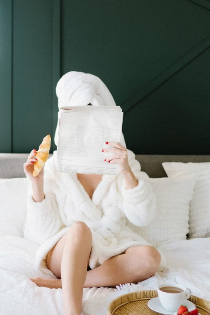 Photo of a woman in a hotel robe and towel on her head, lounging on a bed while reading a newspaper and eating a croissant.