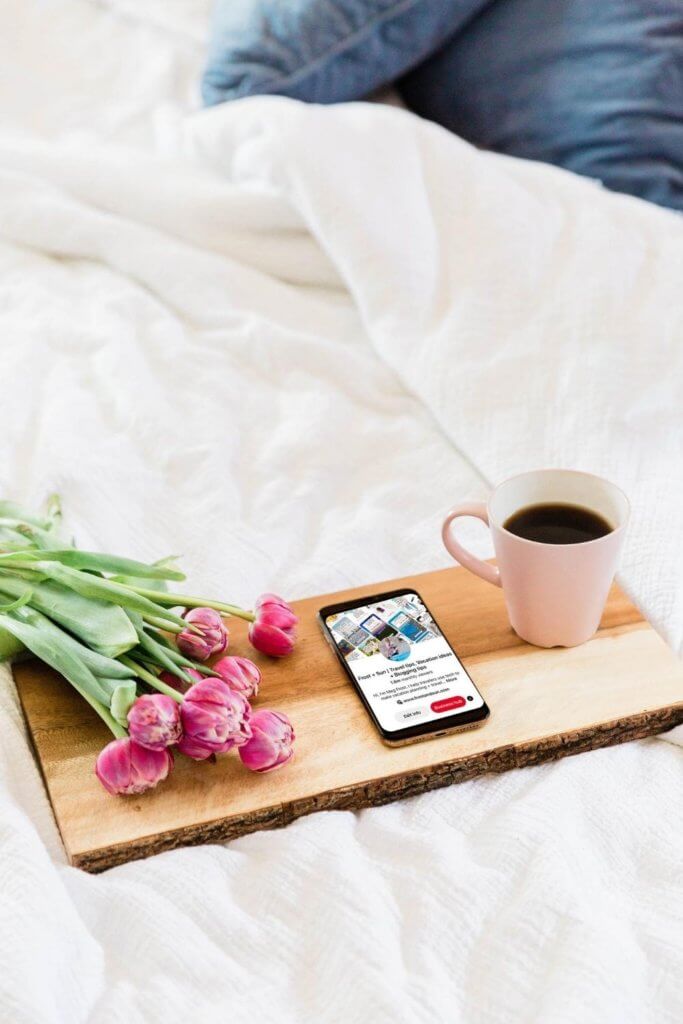 Photo of a wooden tray on an unmade bed. The tray has a bunch of pink flowers, a cup of coffee and a mobile phone open to a website.