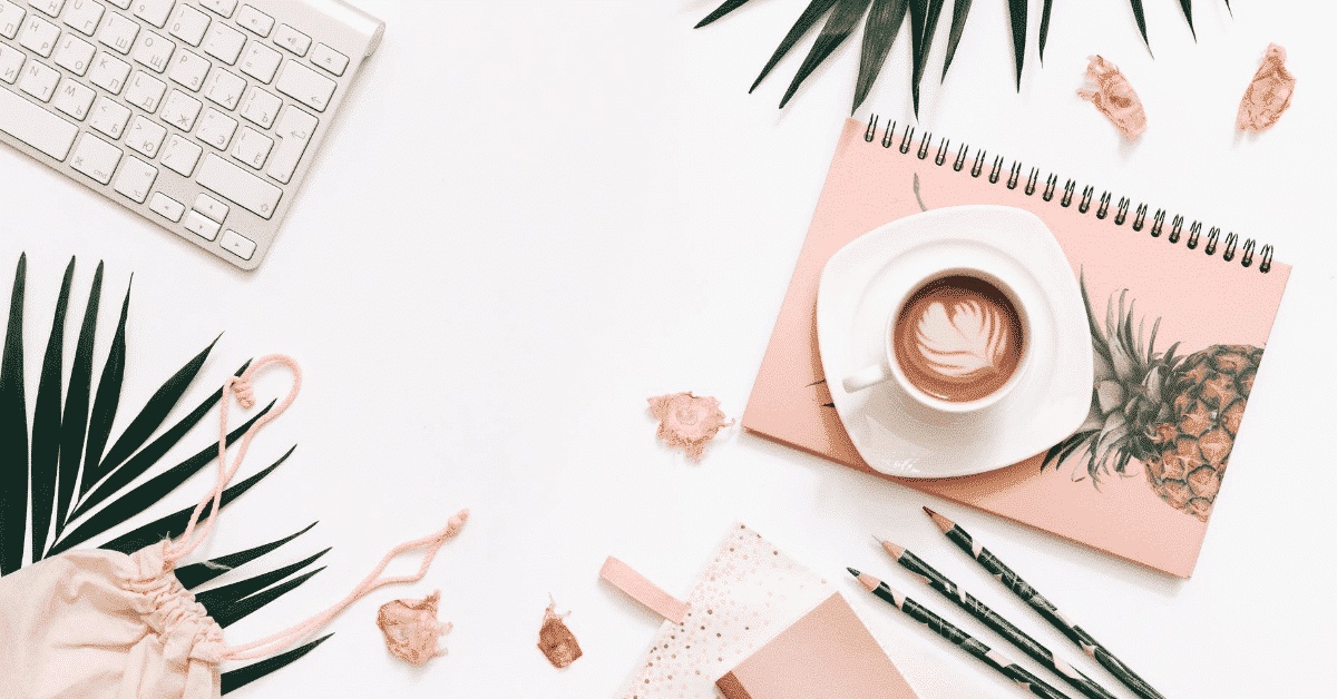 Flat lay photo of a white desk with a latte, green leaves, pencils, a keyboard, and pink notebooks.