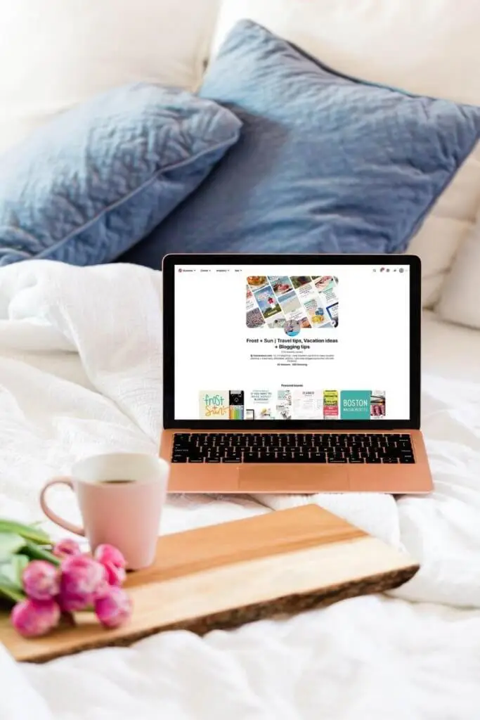 Photo of a laptop open to Pinterest on an unmade bed with white linens. A wooden tray sits nearby with a bunch of pink tulips and a cup of coffee.