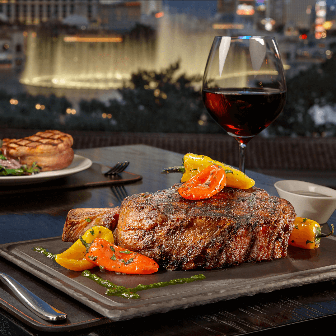 Closeup of a steak dinner with a glass of red wine with the Bellagio Vegas fountains in the background.