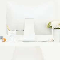 Photo of a desk with a podcast microphone, computer monitor, a vase with white flowers, and several books and journals artfully arranged.