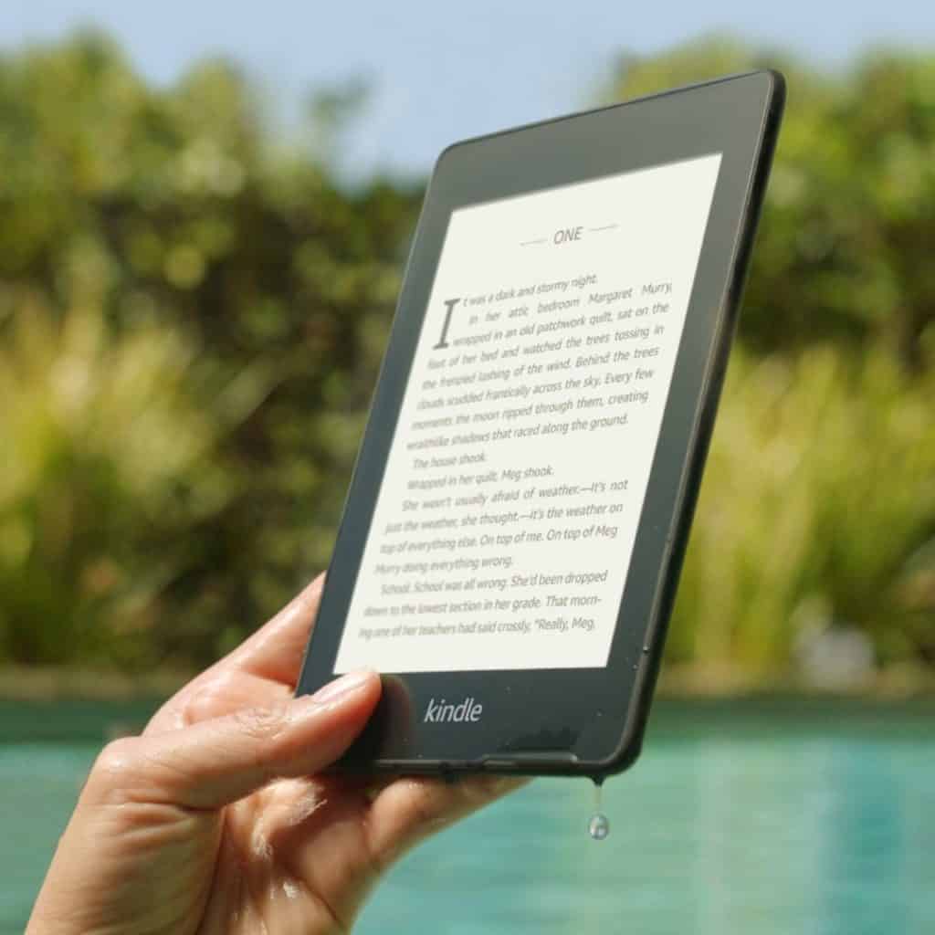 Closeup of a person holding up a Kindle Paperwhite reader with a pool in the background.