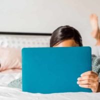 Woman laying across her bed on her stomach with a laptop open, obscuring her face.