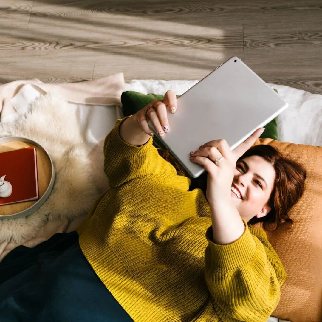 Photo of a woman laying down with her arms extended above her, holding an iPad.