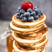 Closeup of a tall stack of thick pancakes with blueberries, a big strawberry, and maple syrup on top.