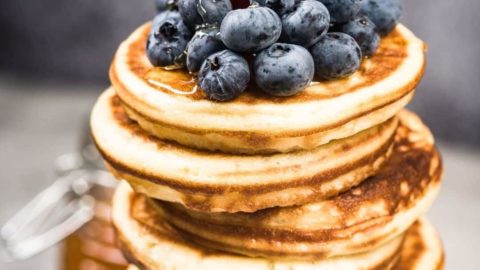 Closeup of a tall stack of thick pancakes with blueberries, a big strawberry, and maple syrup on top.