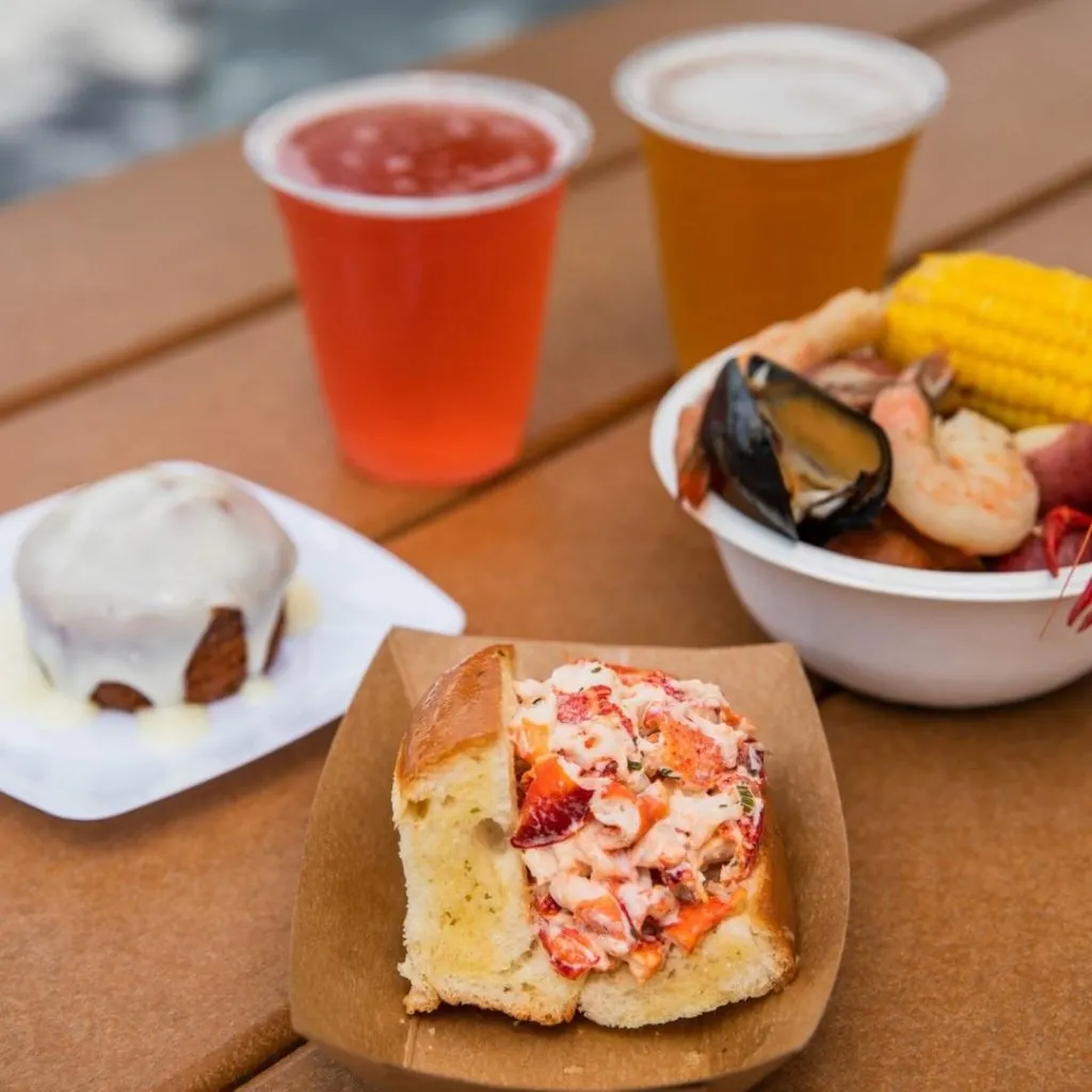 Closeup of 2 beers, a lobster roll, and a small bowl of food from a traditional New England clambake.