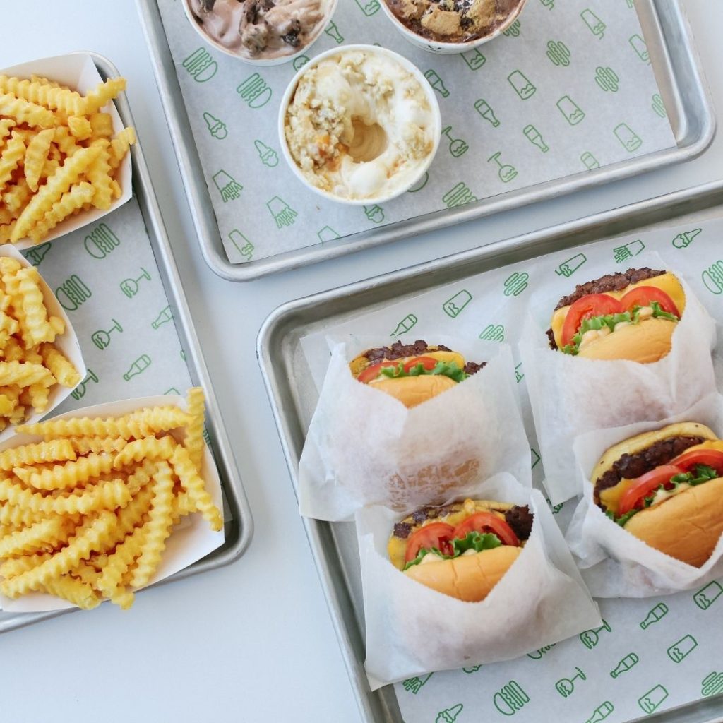 Flat lay photo of 3 dining trays from Shake Shack, one with crinkle french fries, one with milkshakes, and one with cheeseburgers.