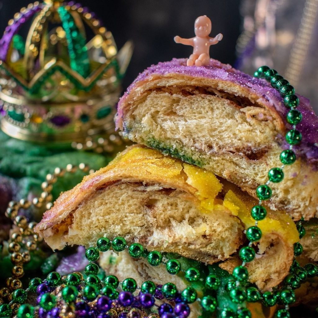 Closeup of sliced king cake with Mardi Gras beads and a plastic baby arranged on top.