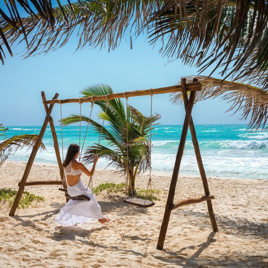 Photo of a woman on a wooden swing on a beach, looking out toward the ocean.