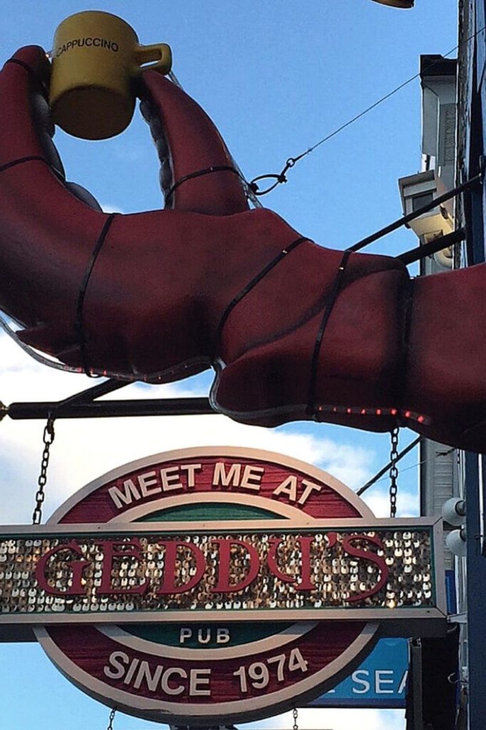 Closeup of the sign for Geddy's. Above is a giant lobster claw holding a yellow cappucino mug.