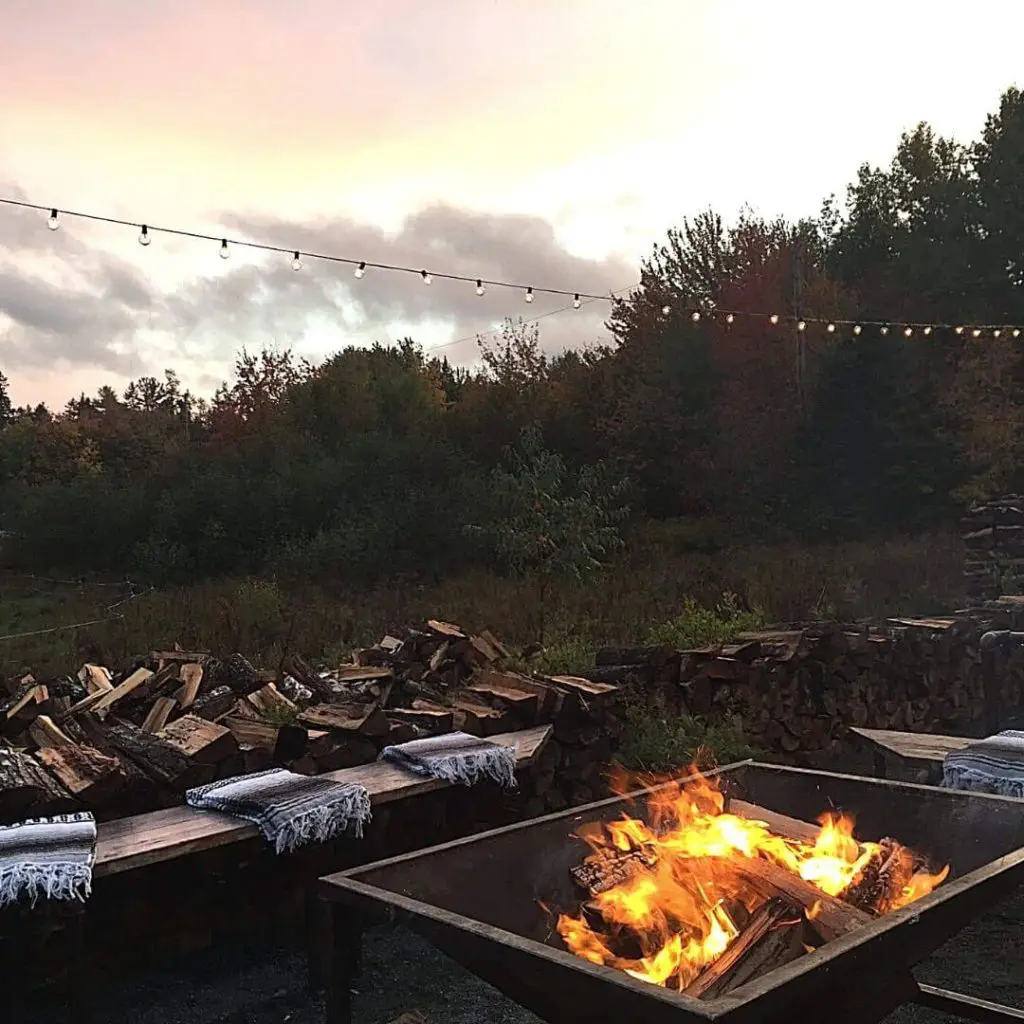 Snapshot of a firepit with benches set around it. In the background are piles of wood, trees, and outdoor string lights.