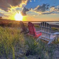 Closeup of 3 empty Adirondack-style chairs sitting on a beach overlooking the sun as it sets over the Atlantic ocean from a Cape Cod beach.