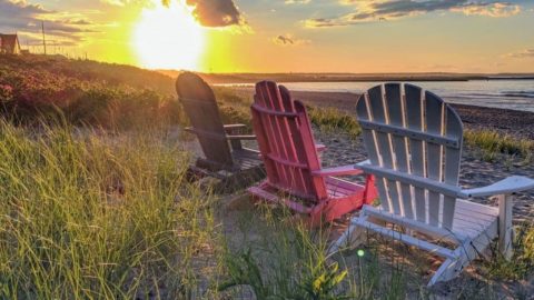 Closeup of 3 empty Adirondack-style chairs sitting on a beach overlooking the sun as it sets over the Atlantic ocean from a Cape Cod beach.