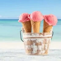 Photo of 3 ice cream cones with bright pink ice cream sitting in a wooden bucket with the beach in the background.