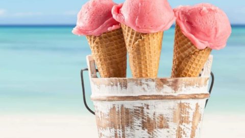 Photo of 3 ice cream cones with bright pink ice cream sitting in a wooden bucket with the beach in the background.