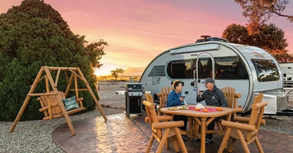 Photo of a couple drinking wine at a picnic table next to a camping trailer.