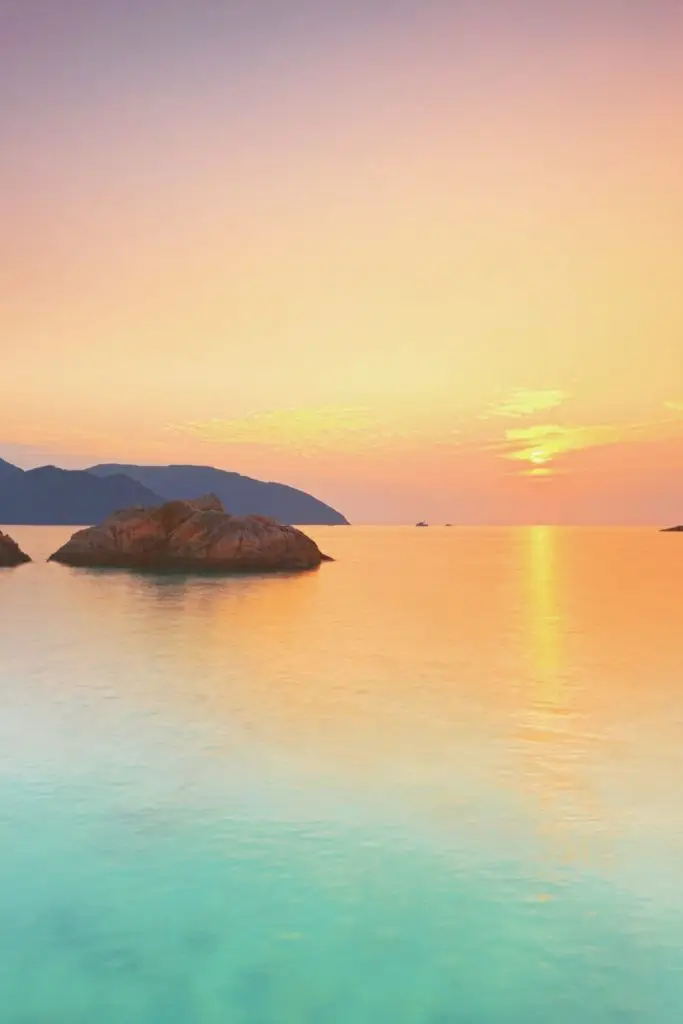 Photo of a candy-colored sunset view with the ocean in the foreground and mountains in the background.