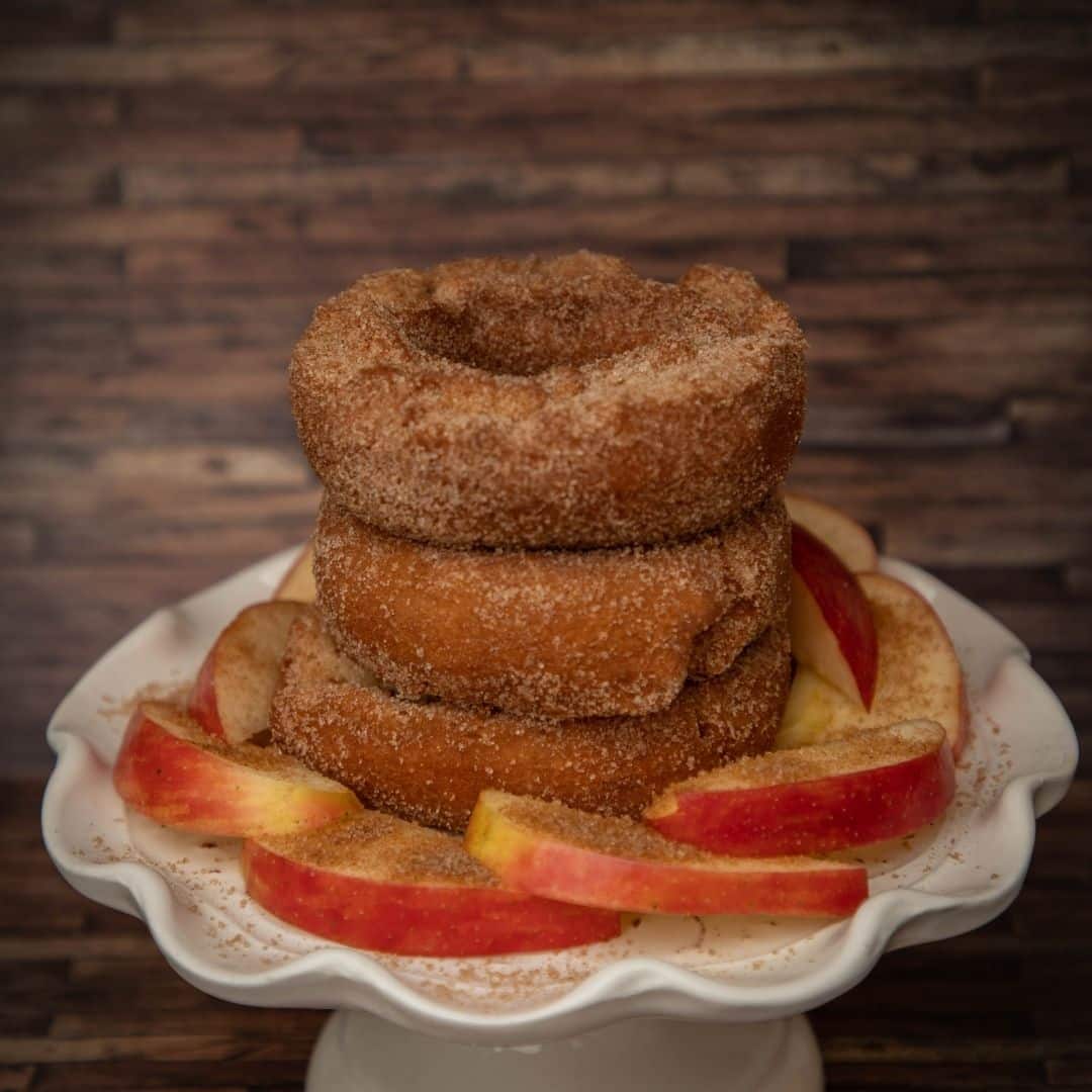 Closeup of a stack of 3 apple cider donuts with apple slices arranged around them on a white pedestal platter.