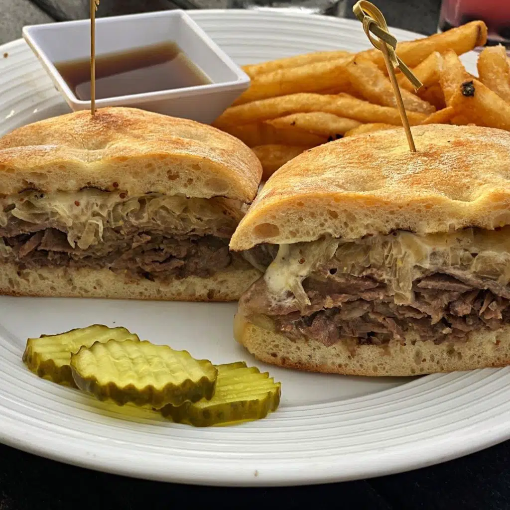 Closeup of the Dublin pub dip sandwich with ribeye beef, carmelized onions, and horseradish cream. Golden crispy fries and aus jus dip are placed behind the sandwich and a stack of pickle chips are in front.