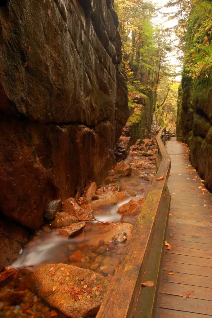 Photo of the wooden walkway along the Flume Gorge Trail in New Hampshire in the Fall with orange and brown leaves on the ground.