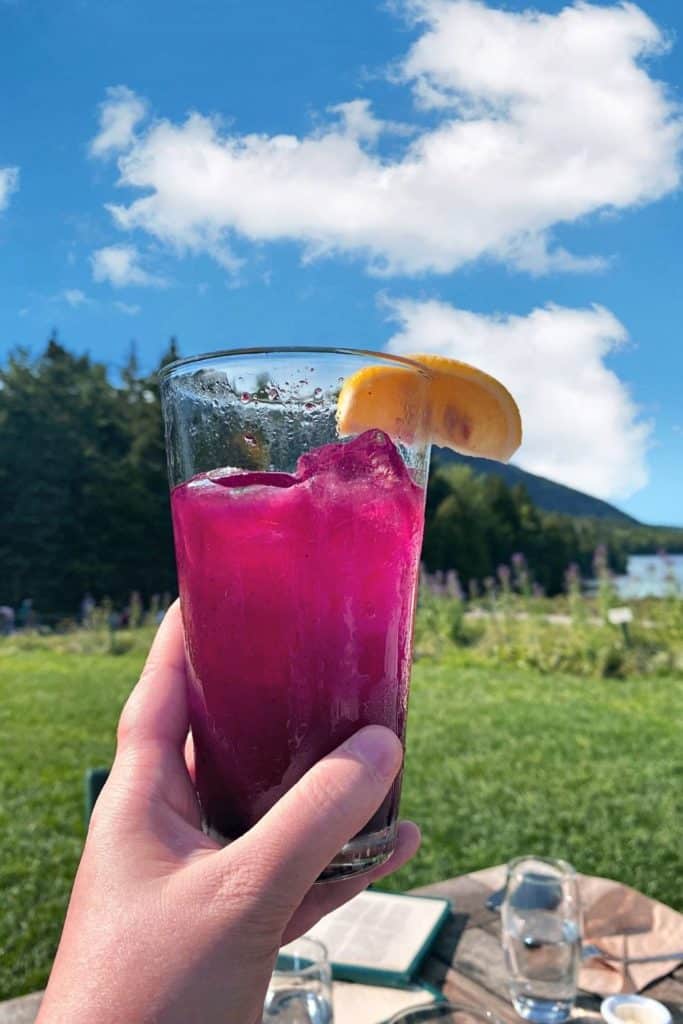 Hand holding a tall glass of blueberry lemonade in front of the Bubbles mountains at Jordan Pond House.