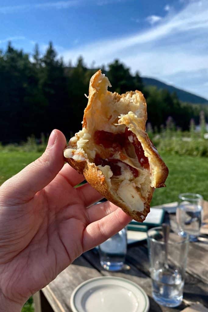 Closeup of a hand holding a popover torn in 2, smeared with butter and strawberry jam with Acadia National Park in the background.