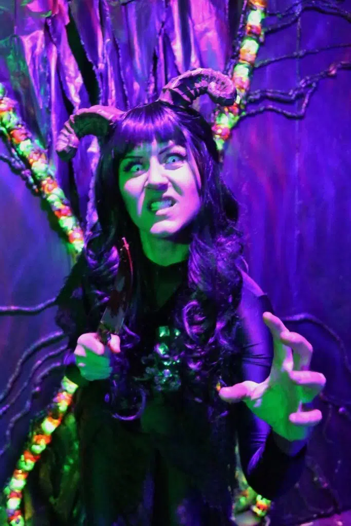 Photo of a woman with horns attached to her head in an intimidating pose. The entire photo is lit up with a purple hue.