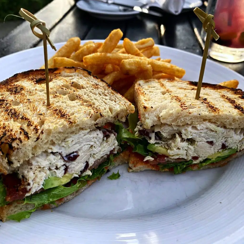 Closeup of a sandwich cut in half and stuffed with rotisserie chicken, avocado, bacon, and lettuce, with crispy golden french fries behind the sandwich.