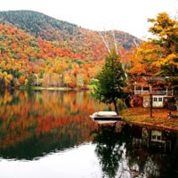 Photo of a lake in Vermont with a tree-lined mountain in the background bursting in Fall foliage colors like orange, yellow, and red.
