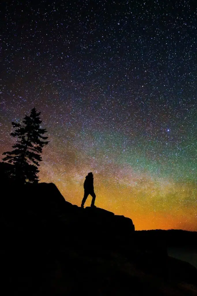 Photo of a silhouette of a man and a tree on a cliff against a starry sky with colors of orange, green, purple, and blue.