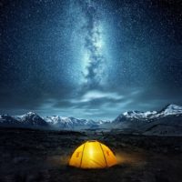 Photo of a yellow tent in the middle of an open field with mountains and a starry sky with the Milky Way galaxy in the background.