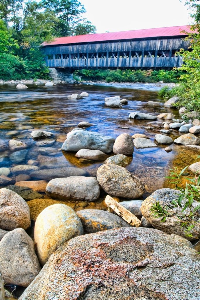 Photo of an old covered bridge in New Hampshire with a creek and rocks in the foreground.