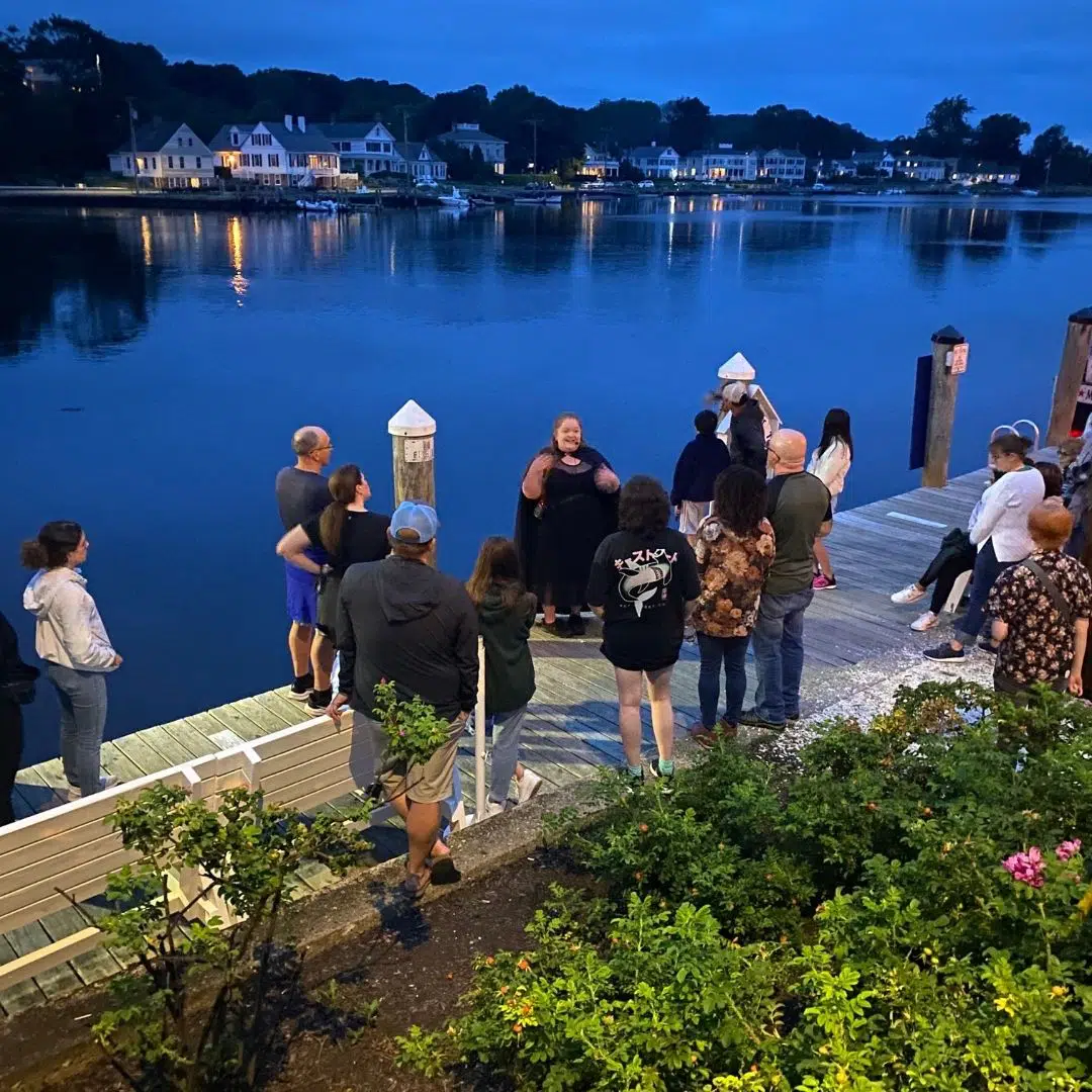 Photo of a group of people gathered on a dock, focused on a woman in a black cape.