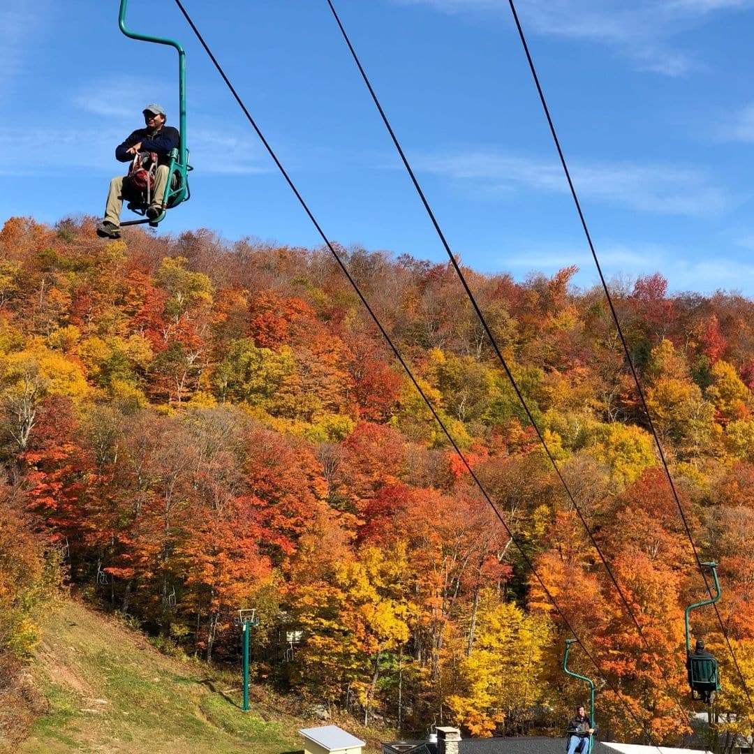 Photo of a man riding a ski lift with Fall foliage in the background.
