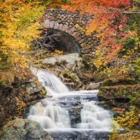 Photo of a stone bridge with a waterfall surrounded by Fall colors.
