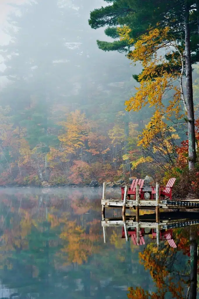 Photo of 2 red Adirondack style chairs sitting on a doc over a lake with Fall foliage surrounding it.