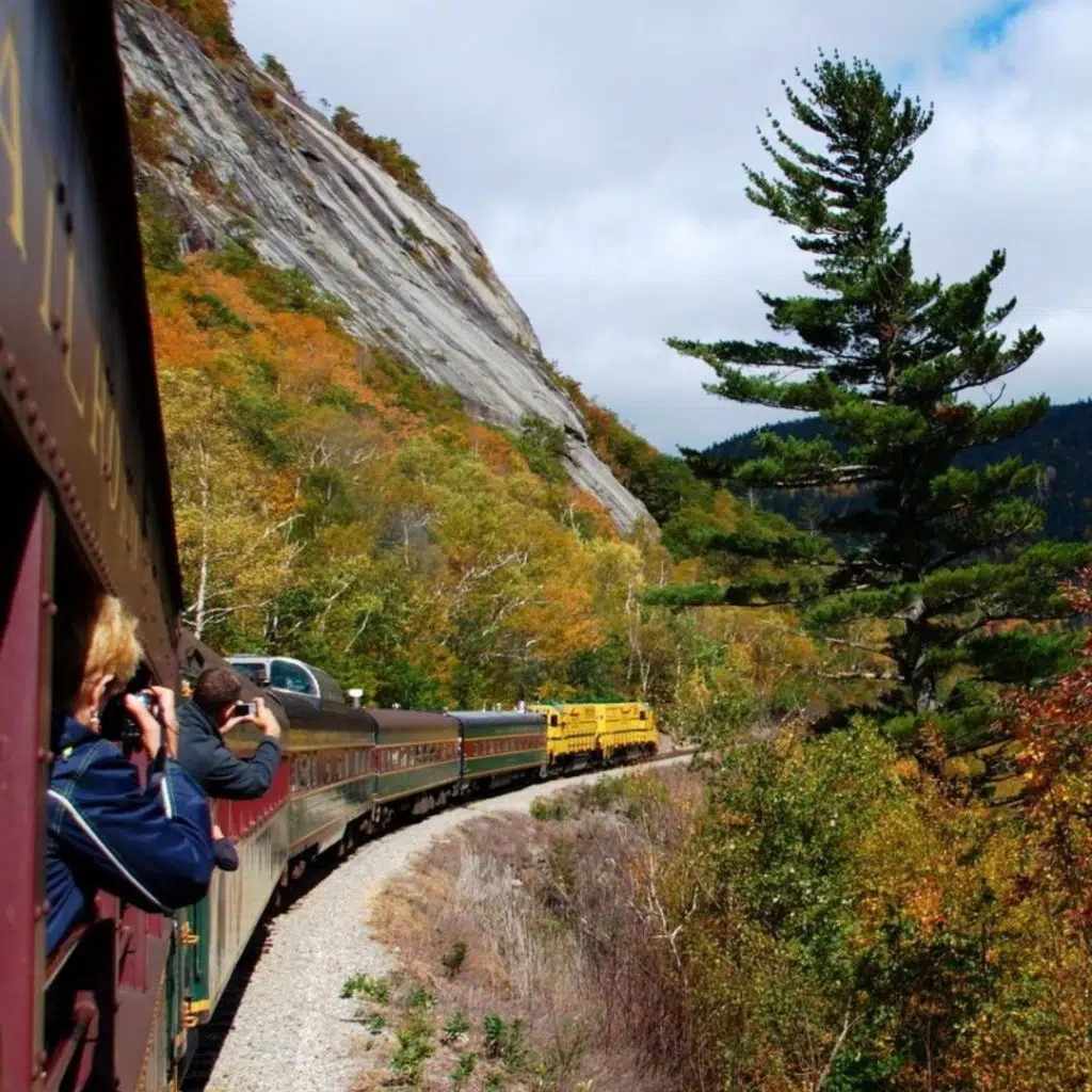 Photo of a train driving past a mountain covered in Fall foliage.