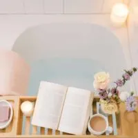 Top down photo of a bathtub filled with water with a tray across the top filled with a book, candle, cup of coffee and flowers.