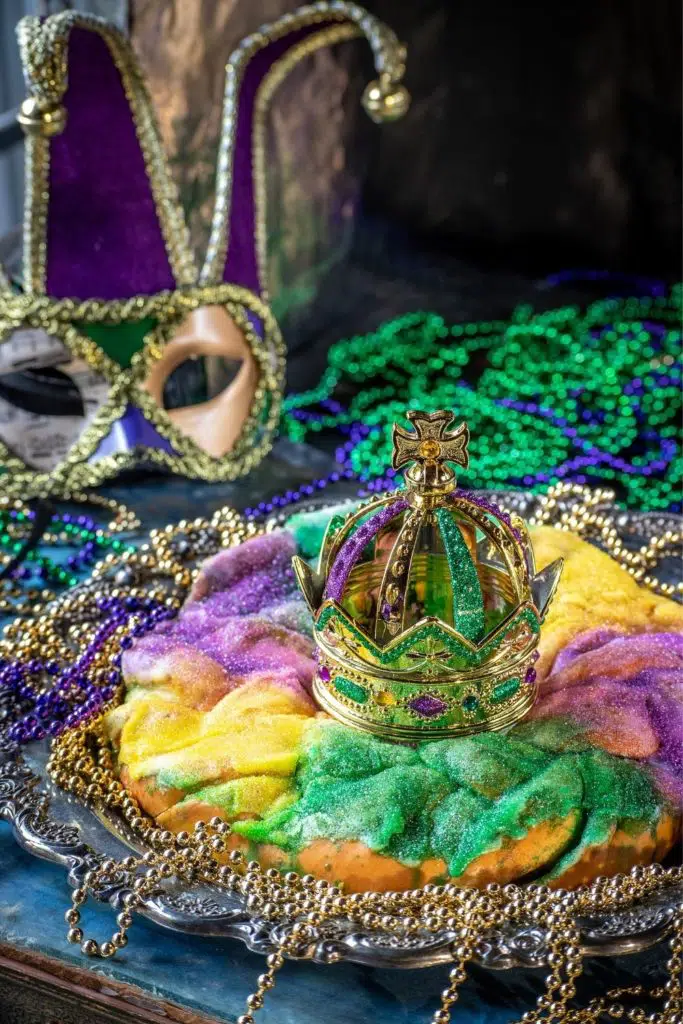 Photo of a Mardi Gras King Cake with a crown in the middle and lots of beads and masks in the background.