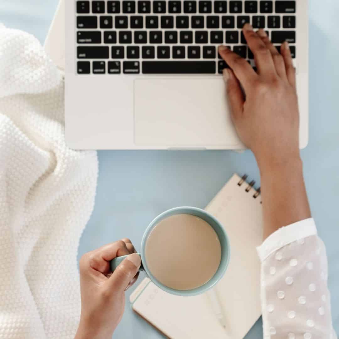 Top-down photo of a woman holding a cup of coffee in her left hand and typing on a laptop with her right hand.