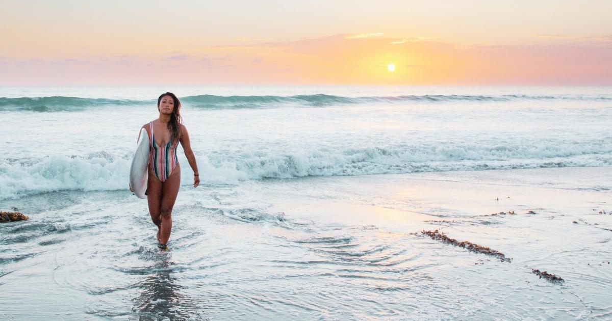 Photo of a woman walking out of the ocean while carrying a surfboard while the sun rises.