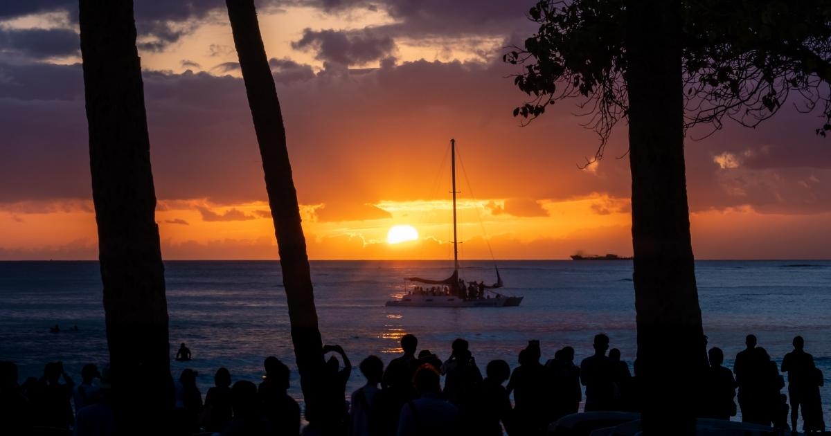Photo of people watching the sunset from Waikiki Beach with a catamaran in the ocean.